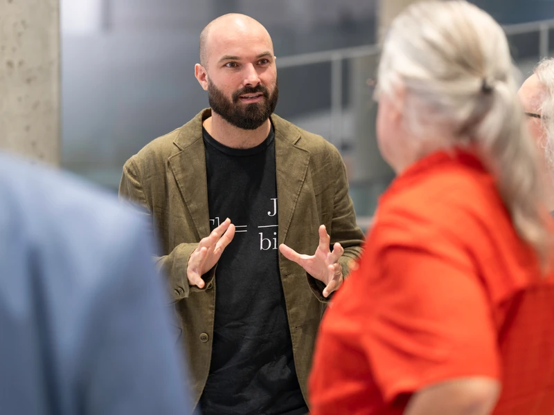 Charles Alexandre Bédard in conversation, gesturing with both hands, wearing his green jacket and J/bit T-shirt.